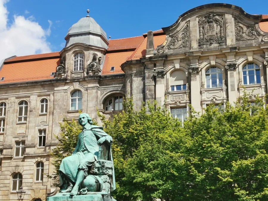 Eine Statue eines sitzenden Mannes in historischer Kleidung vor einem architektonisch beeindruckenden Gebäude mit einem roten Dach und grünen Bäumen.