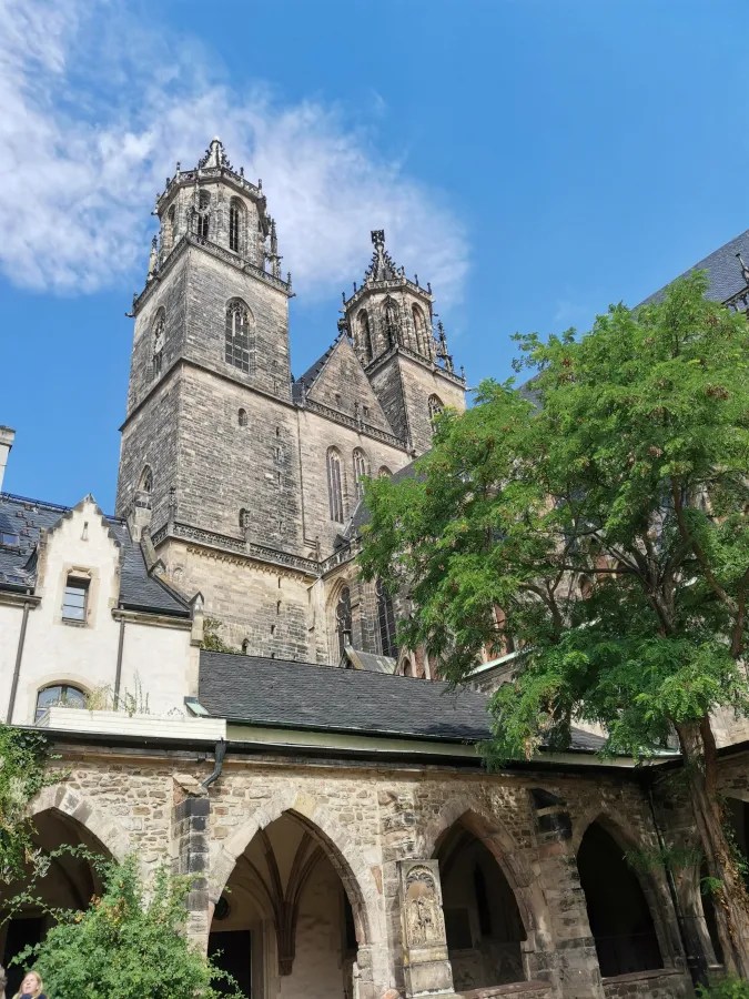 Ansicht einer historischen Kirche mit zwei markanten Türmen und gotischer Architektur, umgeben von Bäumen und einem klaren blauen Himmel.