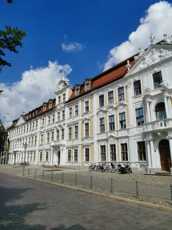 Eine elegante historische Gebäudefassade mit weißen und gelben Elementen, flankiert von Fahrrädern auf einem gepflasterten Weg, unter blauem Himmel mit einigen Wolken.