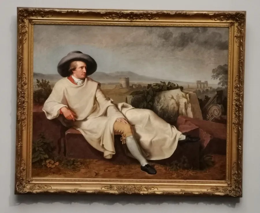 Porträt eines sitzenden Mannes in eleganter Kleidung mit Hut, vor einer Landschaft mit Ruinen im Hintergrund.