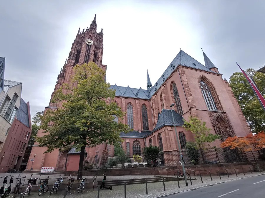 Seitenansicht der roten Kathedrale mit einem hohen Turm und gotischer Architektur, umgeben von Bäumen und einer Straße mit Fahrrädern.