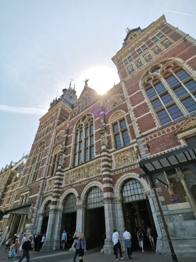Fassade eines historischen, detailreich gestalteten Gebäudes mit großen Fenstern und Statuen, von unten fotografiert, während die Sonne im Hintergrund scheint.