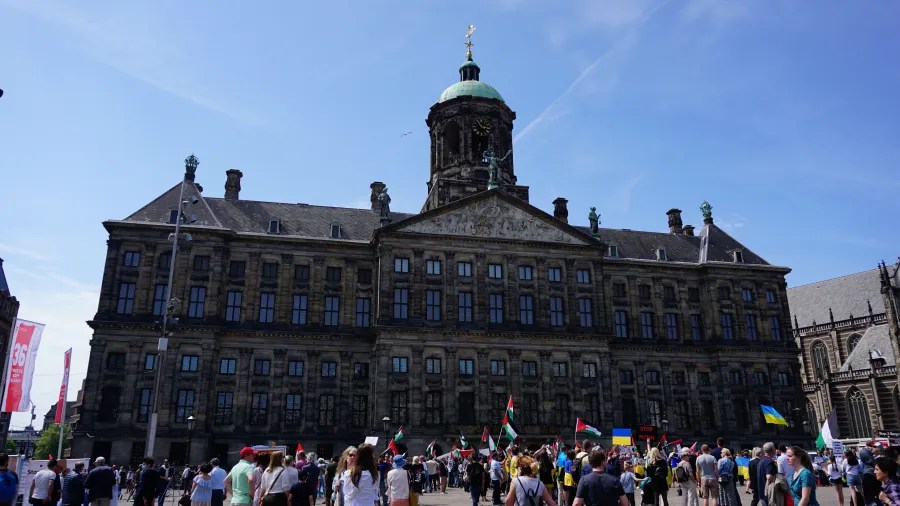 Menschenmenge vor einem historischen Gebäude bei Sonnenschein, mit Flaggen verschiedener Länder.