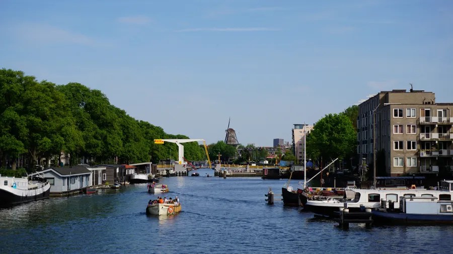 Ein idyllischer Kanal mit Booten und grünen Bäumen entlang der Ufer, im Hintergrund ein Windmühlen-Silhouette und moderne Gebäude.