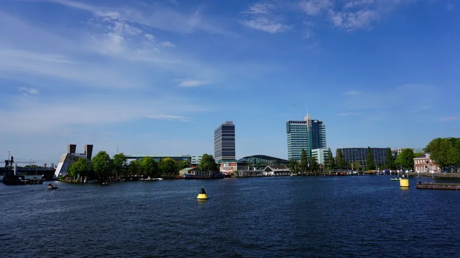 Blick auf einen Stadtfluss mit modernen Gebäuden und Bäumen am Ufer unter einem blauen Himmel.