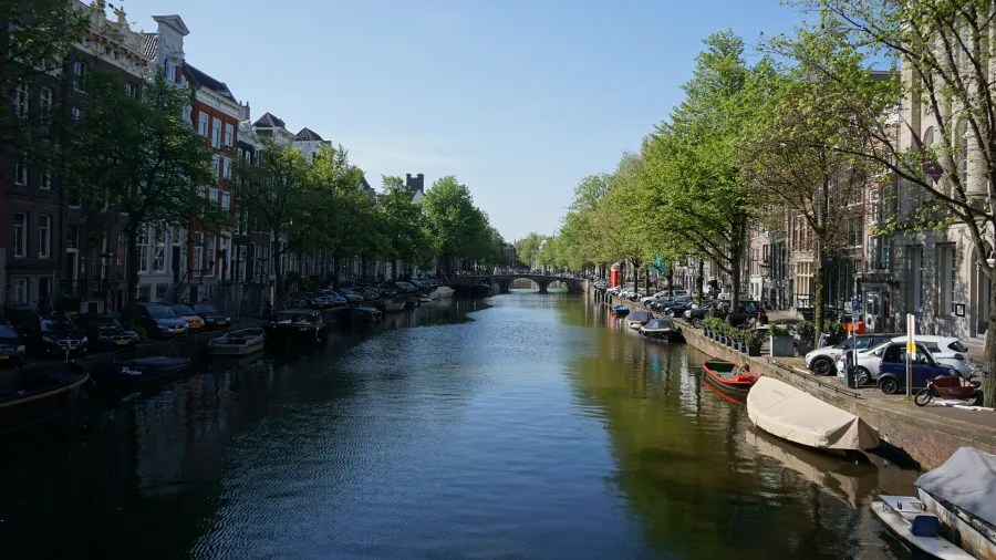 Ein ruhiger Kanal in Amsterdam mit historischen Gebäuden, Bäumen und Booten am Ufer bei klarem Himmel.