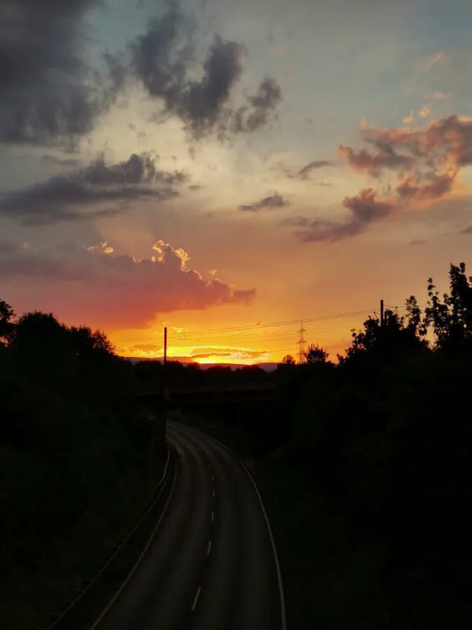 Eine kurvenreiche Straße führt in den Sonnenuntergang, mit dramatischen Wolken und einem warmen orangefarbenen Himmel.