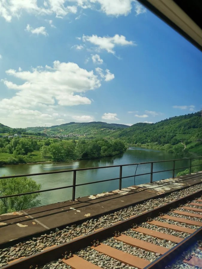Blick aus dem Zugfenster auf eine grüne Landschaft mit einem Fluss, umgeben von sanften Hügeln und einem blauen Himmel mit einigen Wolken.