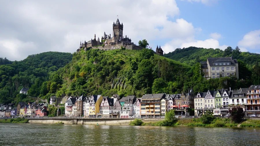 Blick auf die mittelalterliche Burg Metternich, die auf einem Hügel über dem Mosel-Ufer thront, mit bunten Häusern am Wasser.
