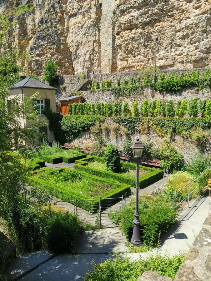 Blick auf einen gepflegten Garten mit geometrischen Hecken, umgeben von einer Felswand und Bänken. Im Hintergrund steht ein kleines Gebäude mit Fenster.