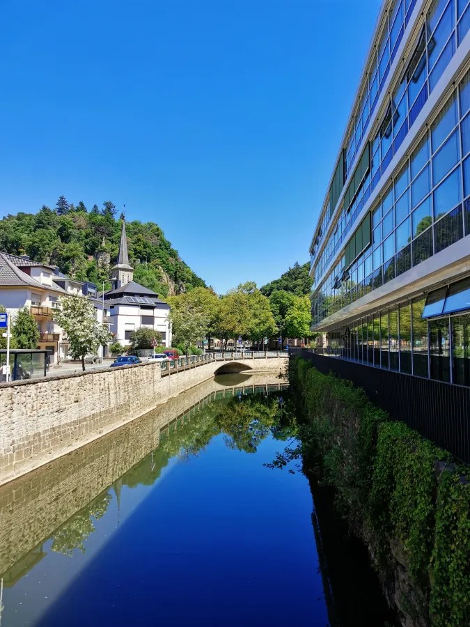 Ein ruhiger Fluss mit Spiegelung der umliegenden Gebäude und Bäume, an einer modernen Glasfassade entlang einer promenadenartigen Uferstraße.