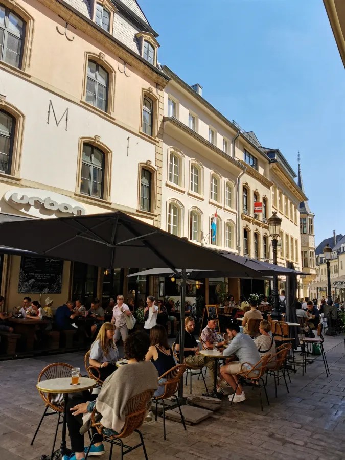 Belebte Straßenszene mit einem Café im Freien, umgeben von historischen Gebäuden und strahlend blauem Himmel.