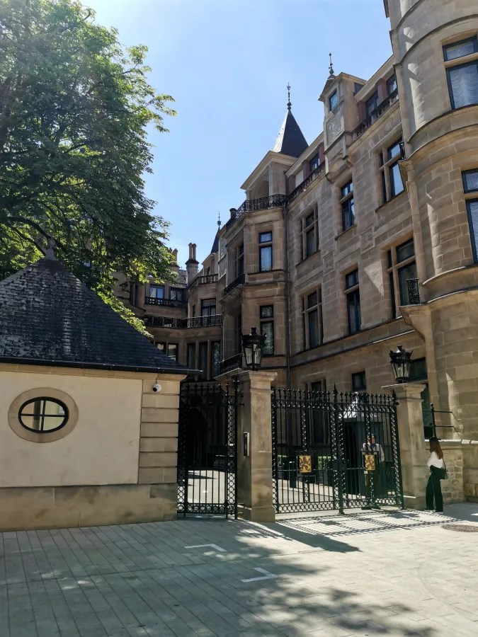 Eingang zu einem eleganten Gebäude mit historischen Architekturdetails und einem Tor, umgeben von Bäumen und Sonnenschein.