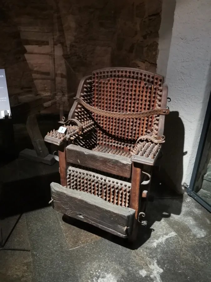 Ein historischer Stuhl mit spitzen Nägeln und Holzrahmen, der in einem Ausstellungsraum präsentiert wird.