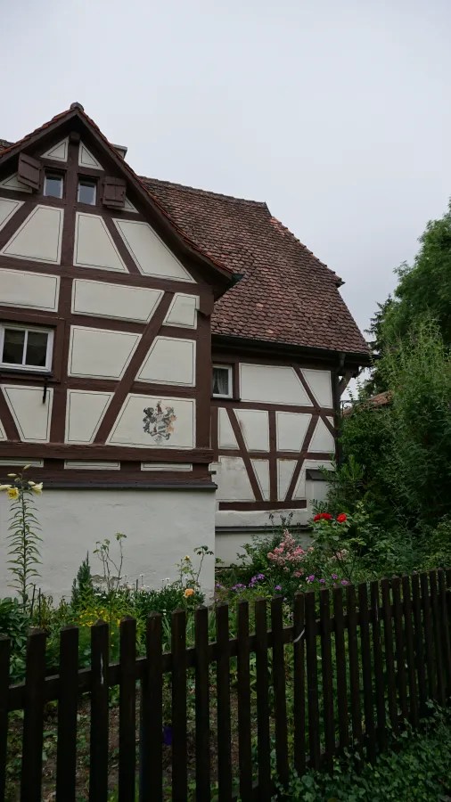 Ansicht eines Fachwerkhauses mit blühendem Garten und Holzzaun, unter einem bewölkten Himmel.
