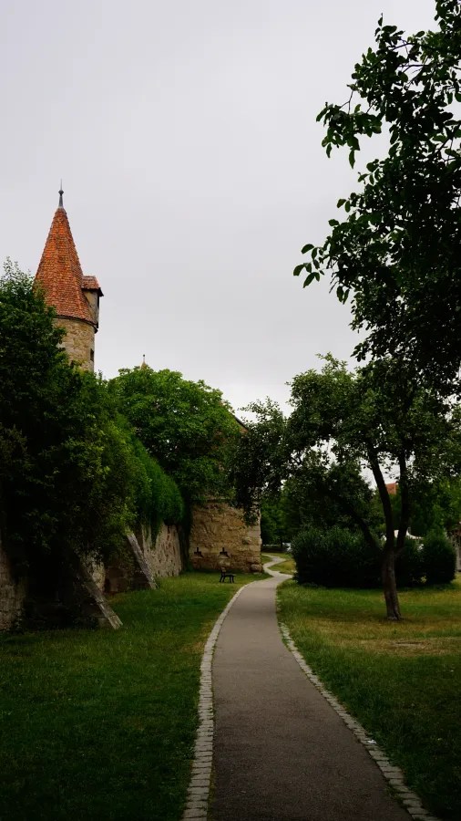 Ein schmaler Weg, der durch eine grüne Landschaft mit Bäumen führt, neben einer alten Stadtmauer und einem Turm mit rotem Dach, unter einem bewölkten Himmel.
