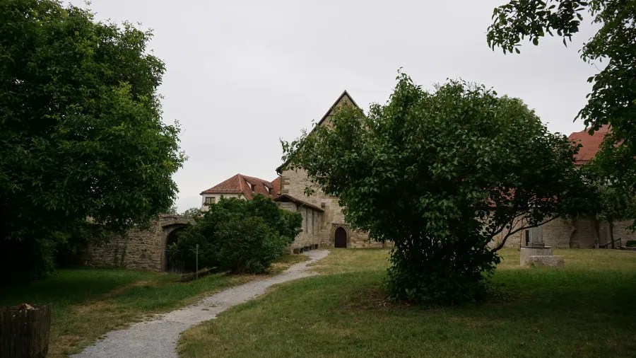 Eine malerische Ansicht eines alten, steinernen Gebäudes, umgeben von Bäumen und einem gepflegten Grünbereich, mit einem schmalen Weg, der in die Szenerie führt.