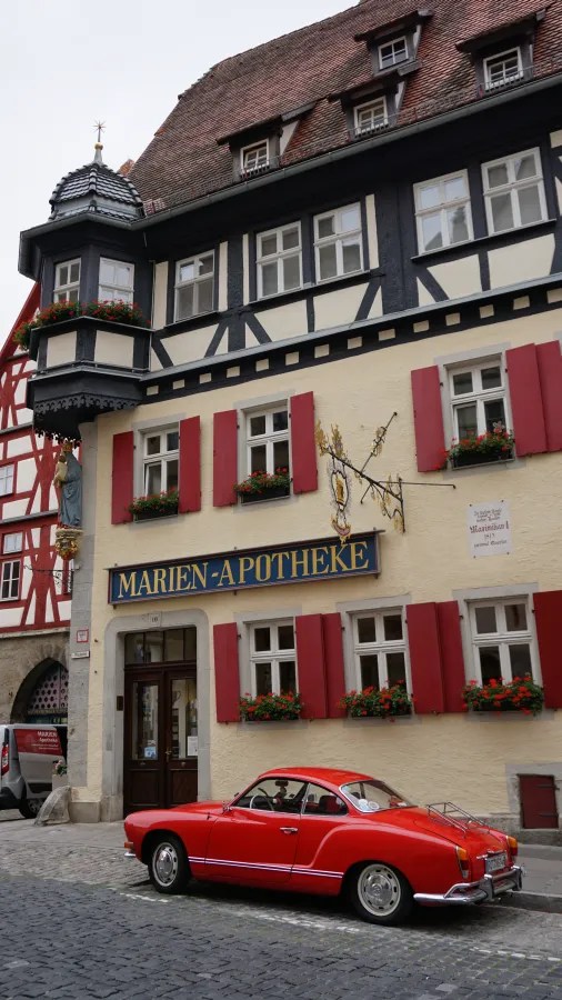 Ein rotes Auto steht vor einem historischen Gebäude mit der Aufschrift 'Marien-Apotheke' und blühenden Blumenkästen an den Fenstern.