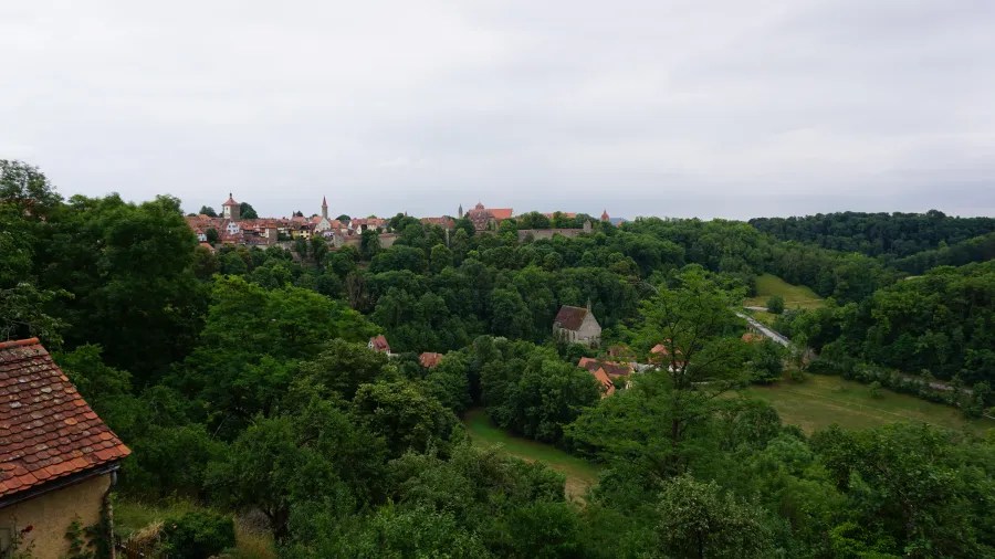 Panorama einer malerischen Landschaft mit einem Dorf, umgeben von grünen Wäldern und Hügeln unter einem bewölkten Himmel.