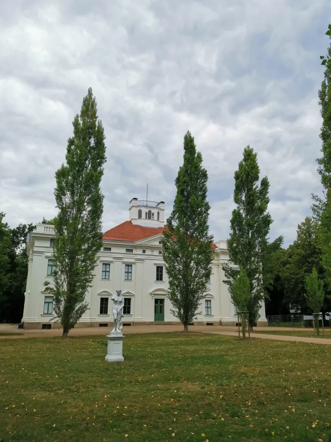 Ein historisches Gebäude mit weißer Fassade und rotem Dach, flankiert von hohen Bäumen und einer Skulptur in einem Park.