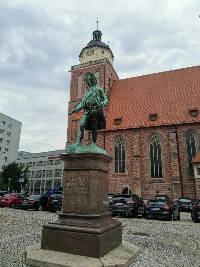 Bronzestatue von Leopold Prinz von Anhalt, steht vor einer Kirche mit rotem Ziegeldach und einem historischen Turm.