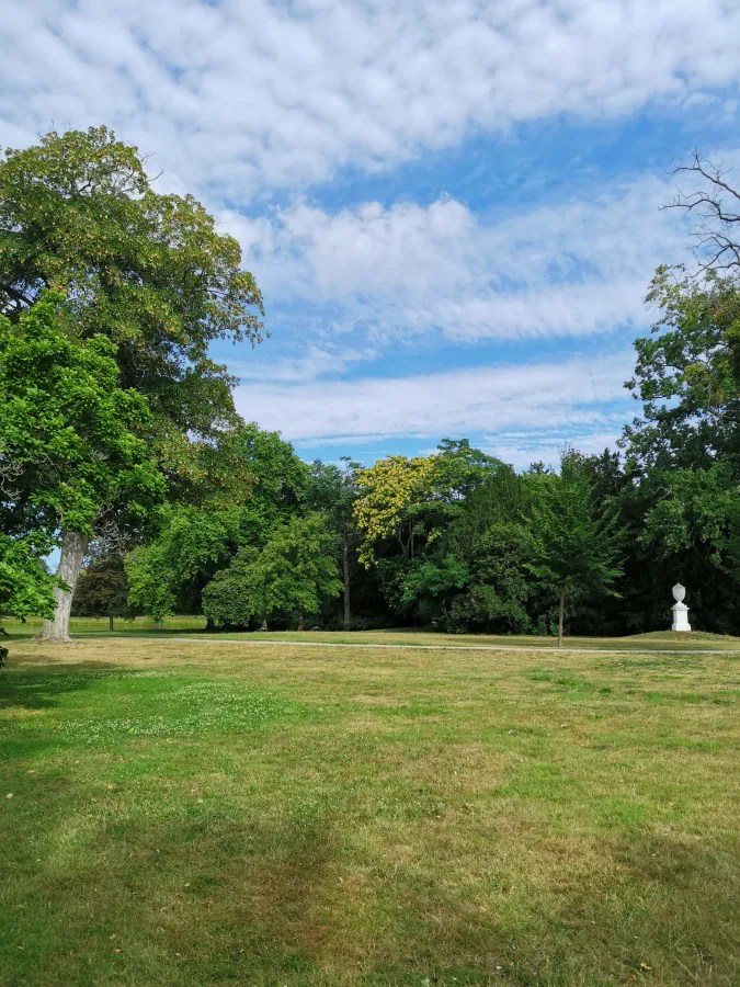 Ein weitläufiger Park mit grünen Wiesen, Bäumen und einem klaren Himmel mit einigen Wolken. Im Hintergrund steht eine Skulptur.