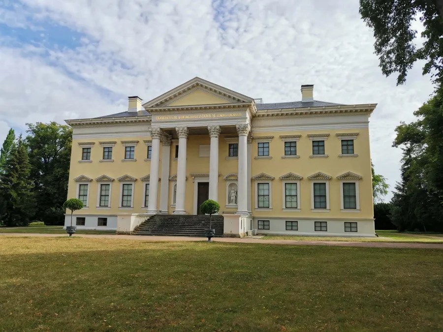 Vorderansicht eines historischen gelben Gebäudes mit Säulen und Stufen, umgeben von Grünflächen und Bäumen unter einem bewölkten Himmel.