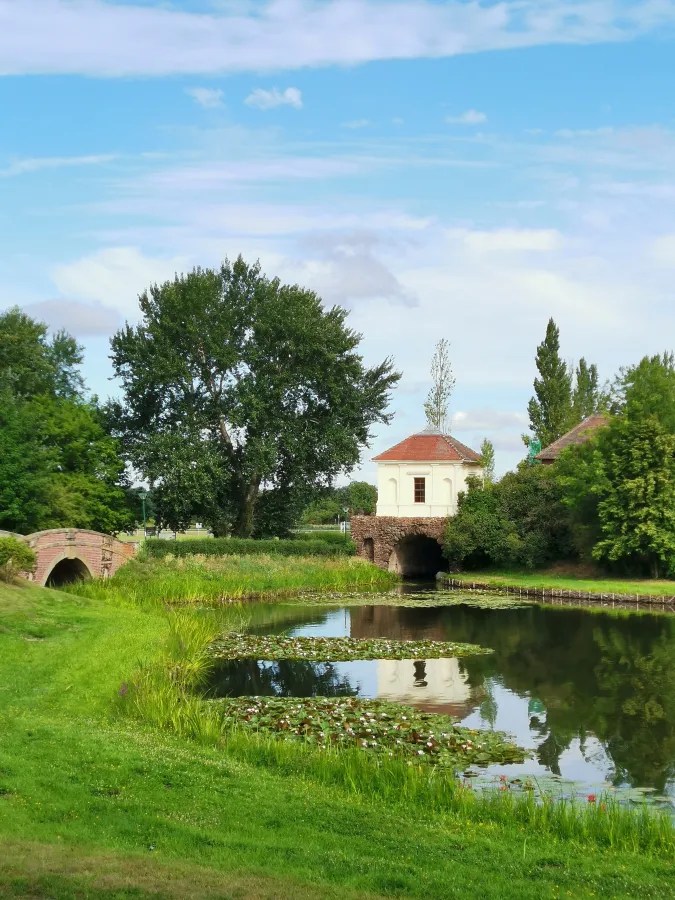 Ein ruhiger Park mit einem Fluss und einer kleinen Brücke, umgeben von Bäumen und üppigem Grün. Im Hintergrund ist ein historisches Gebäude mit einem roten Dach und einem Reflektor im Wasser sichtbar.