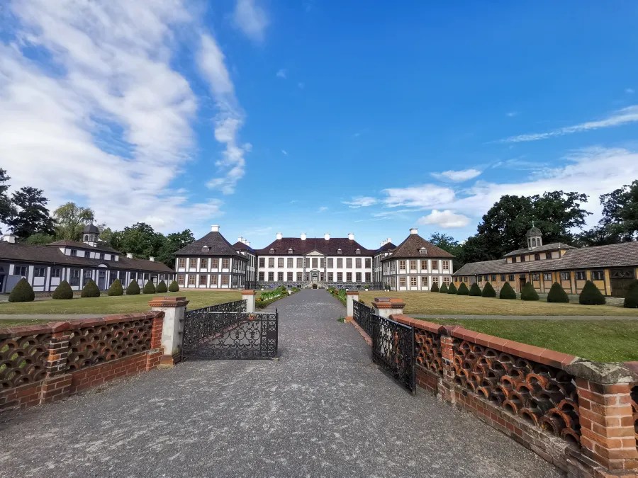 Ein Blick auf ein historisches Herrenhaus mit einem gepflegten Vorplatz, umgeben von Bäumen und einer blauen Himmel.
