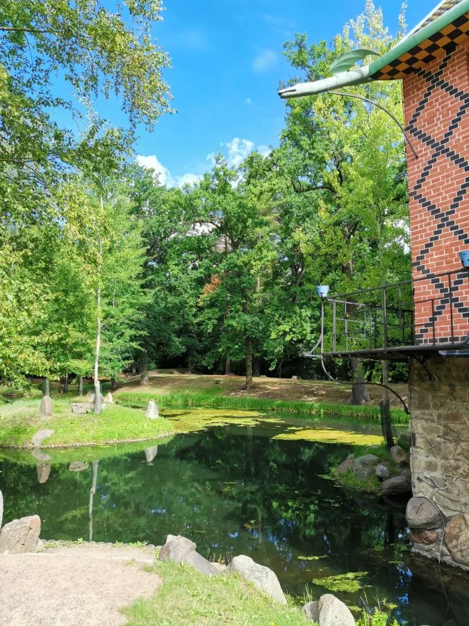 Ein ruhiger Teich umgeben von Bäumen und Felsen, mit einem Gebäude auf der rechten Seite.