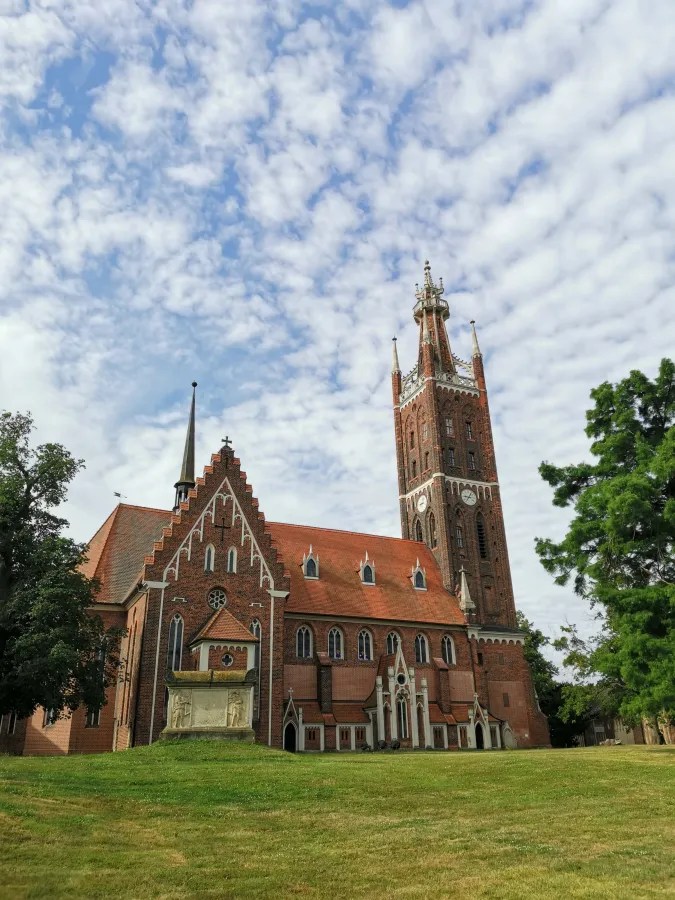 Eine gotische Kirche mit einem hohen Glockenturm, umgeben von begrünter Fläche und einem blauen Himmel mit Wolken.