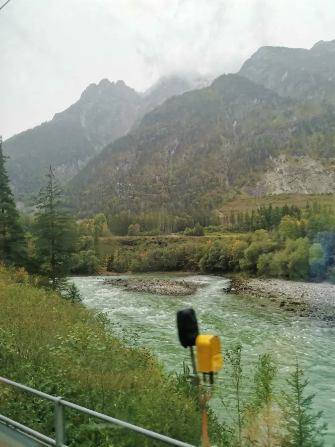 Blick auf einen Fluss, der durch eine grüne Landschaft mit bewaldeten Hügeln und Bergen fließt. Der Himmel ist bewölkt und die Umgebung wirkt herbstlich.