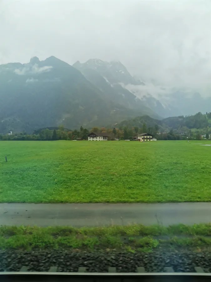 Landschaft mit grünen Wiesen und Bergen im Nebel, in der Nähe von Gebäuden.