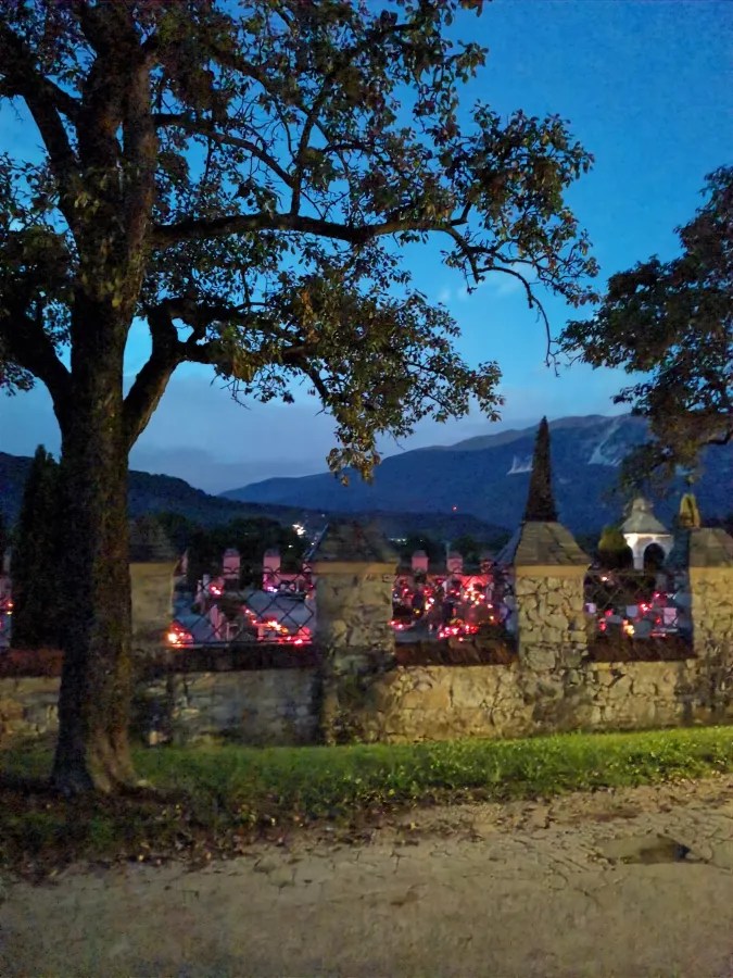 Ein nächtlicher Blick auf einen Friedhof mit leuchtenden Grablichtern, umgeben von einer Steinmauer und Bäumen im Vordergrund.