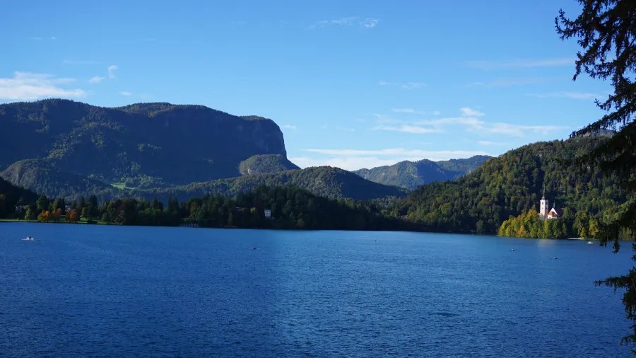 Eine Panoramaansicht eines klaren blauen Sees, umgeben von grünen Hügeln und Bergen, mit einer kleinen Kirche am Ufer.