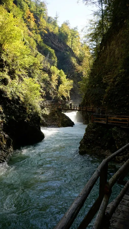 Ein malerisches Tal mit einem klaren, fließenden Wasserlauf und einer Holzbrücke, umgeben von grünen Bäumen und steilen Felsen.