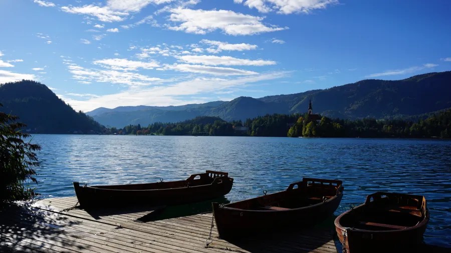 Ruhiger See mit mehreren Booten am Ufer und malerischer Berglandschaft im Hintergrund unter blauem Himmel.