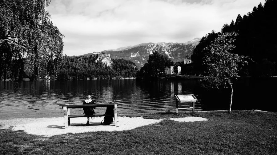 Eine Person sitzt auf einer Bank am Ufer eines ruhigen Sees, umgeben von Bergen und Bäumen. Es ist eine schwarz-weiß Aufnahme.