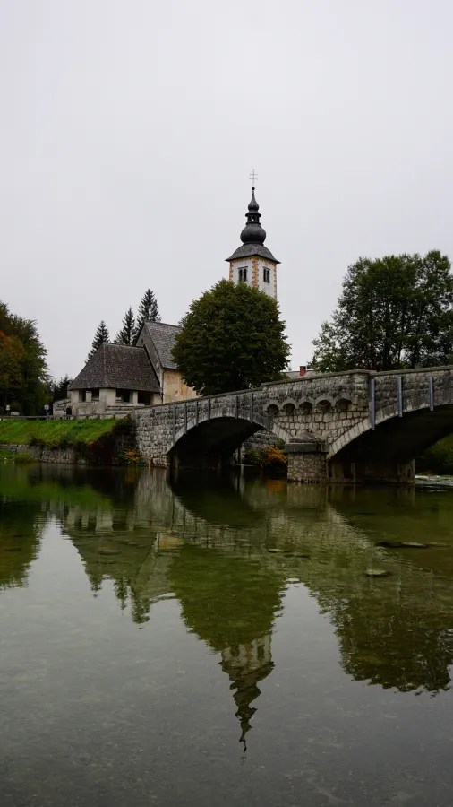Eine malerische Brücke über einen ruhigen Fluss mit dem Spiegelbild eines Kirchturms und Bäumen im Hintergrund.