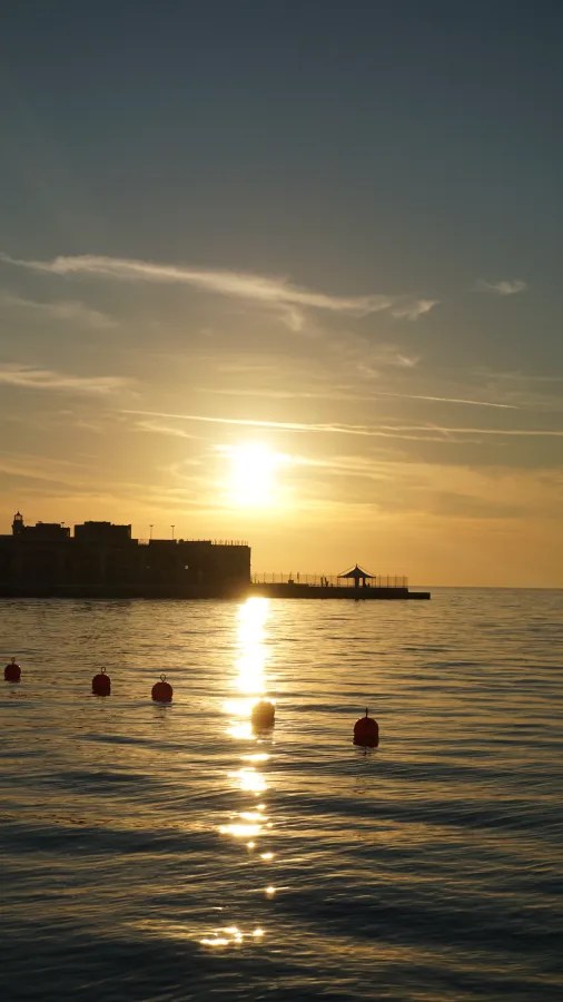 Sonnenuntergang über ruhigem Wasser mit einem Pier und schwimmenden Bojen im Vordergrund.