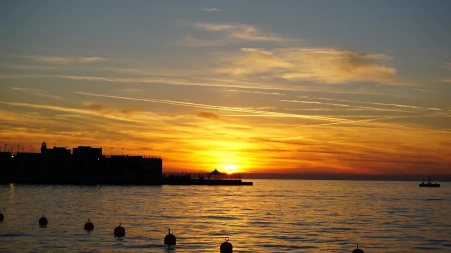 Sonnenuntergang über dem Wasser mit orangefarbenem Himmel und Silhouette eines Anlegers im Hintergrund.