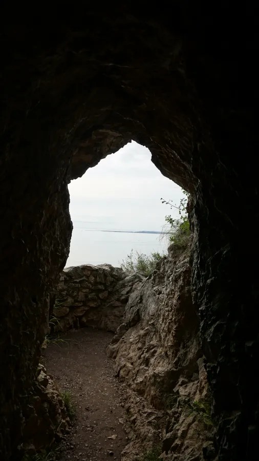 Blick durch einen natürlichen Felsenbogen auf einen ruhigen See und den Himmel, umgeben von grünem Bewuchs.