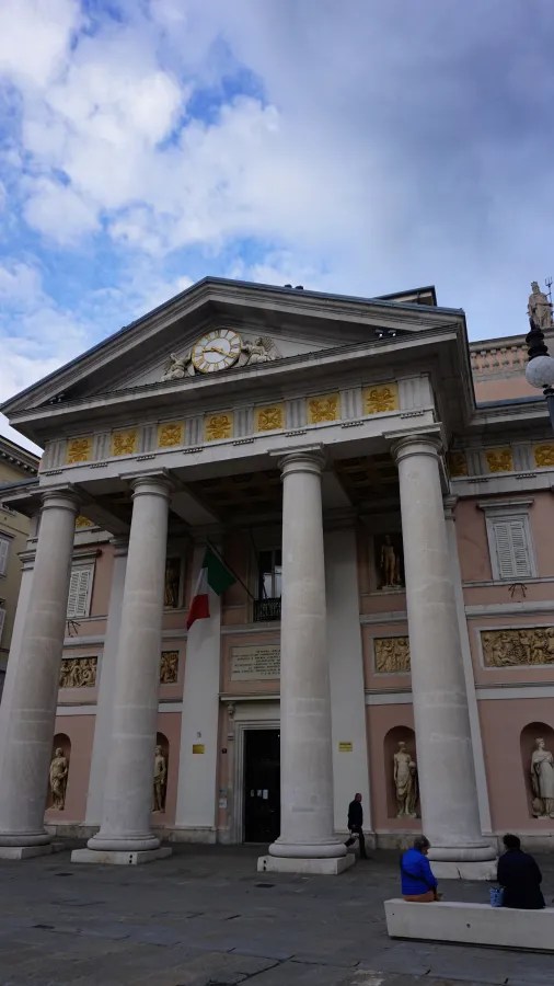 Fassade eines historischen Gebäudes mit Säulen, einer Uhr und Skulpturen, umgeben von einem blauen Himmel.