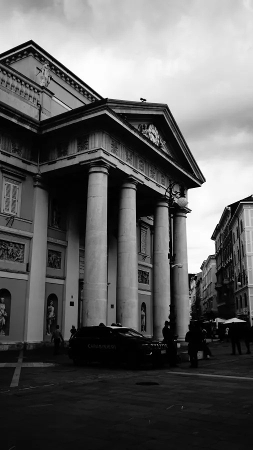 Historisches Gebäude mit Säulen und Skulpturen in der Fassade, unter einem bewölkten Himmel. Im Vordergrund ist ein Polizei-Auto und einige Personen zu sehen.