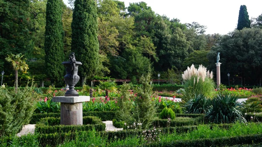 Schöner Garten mit bunten Blumen, hohen Bäumen und einem zentralen Ornament in Form eines Vasenbrunnens.