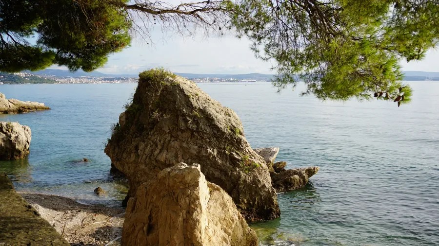 Felsen am Ufer eines ruhigen Gewässers mit Blick auf die Küste und den Himmel, umgeben von Bäumen.