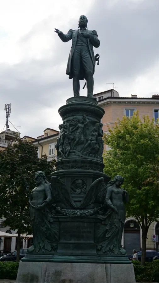 Bronzestatue eines Mannes in Anzug, der auf einem hohen Sockel steht, umgeben von künstlerischen Verzierungen und Engelsskulpturen, unter einem bewölkten Himmel.