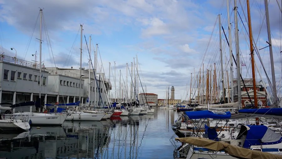 Blick auf einen Hafen mit mehreren Yachten und Segelbooten, umgeben von moderner Architektur und einem Leuchtturm im Hintergrund.