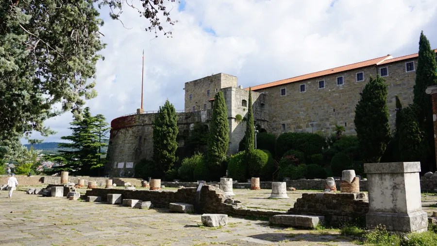Blick auf eine alte Festung mit grünen Gärten und Ruinen in der Vordergrund, unter einem bewölkten Himmel.