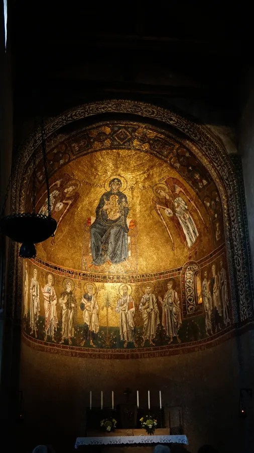 Mosaik mit der Darstellung der Jungfrau Maria, die auf einem Thron sitzt, umgeben von Engeln und Heiligen in einer lichtdurchfluteten Kirche.